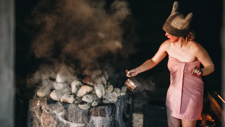 A woman in a horned felt sauna hat throws water on the stones of an Estonian smoke sauna
