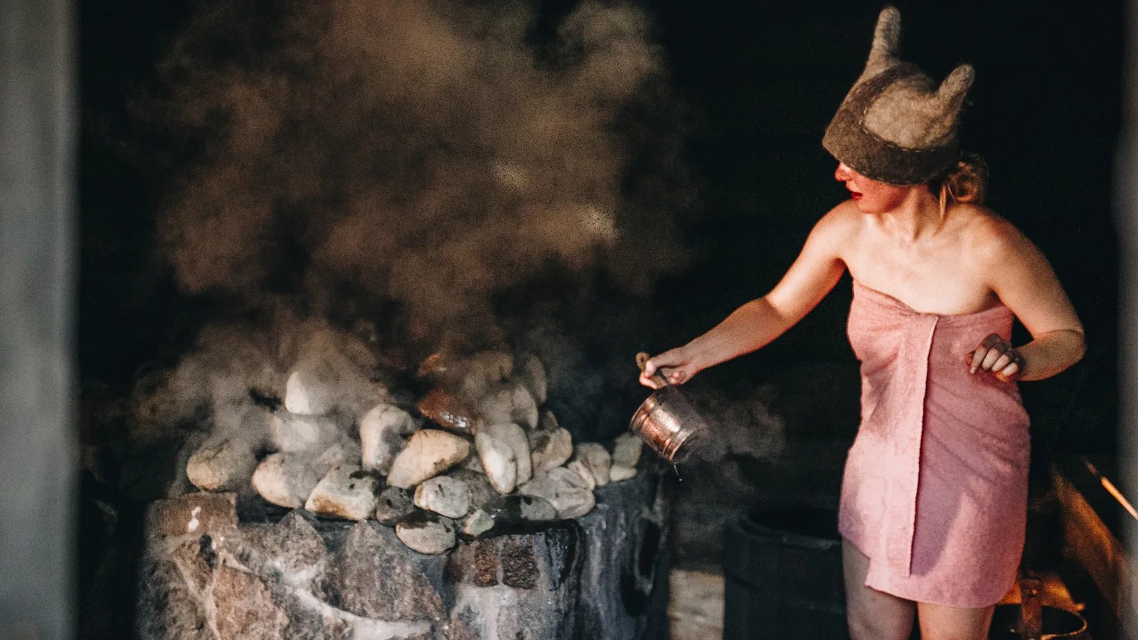 A woman in a horned felt sauna hat throws water on the stones of an Estonian smoke sauna