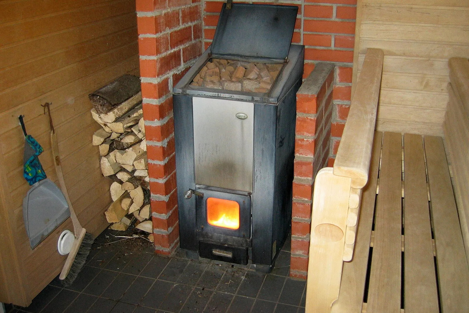 A wood-burning sauna stove with a fire in the firebox and split birch logs stacked beside it