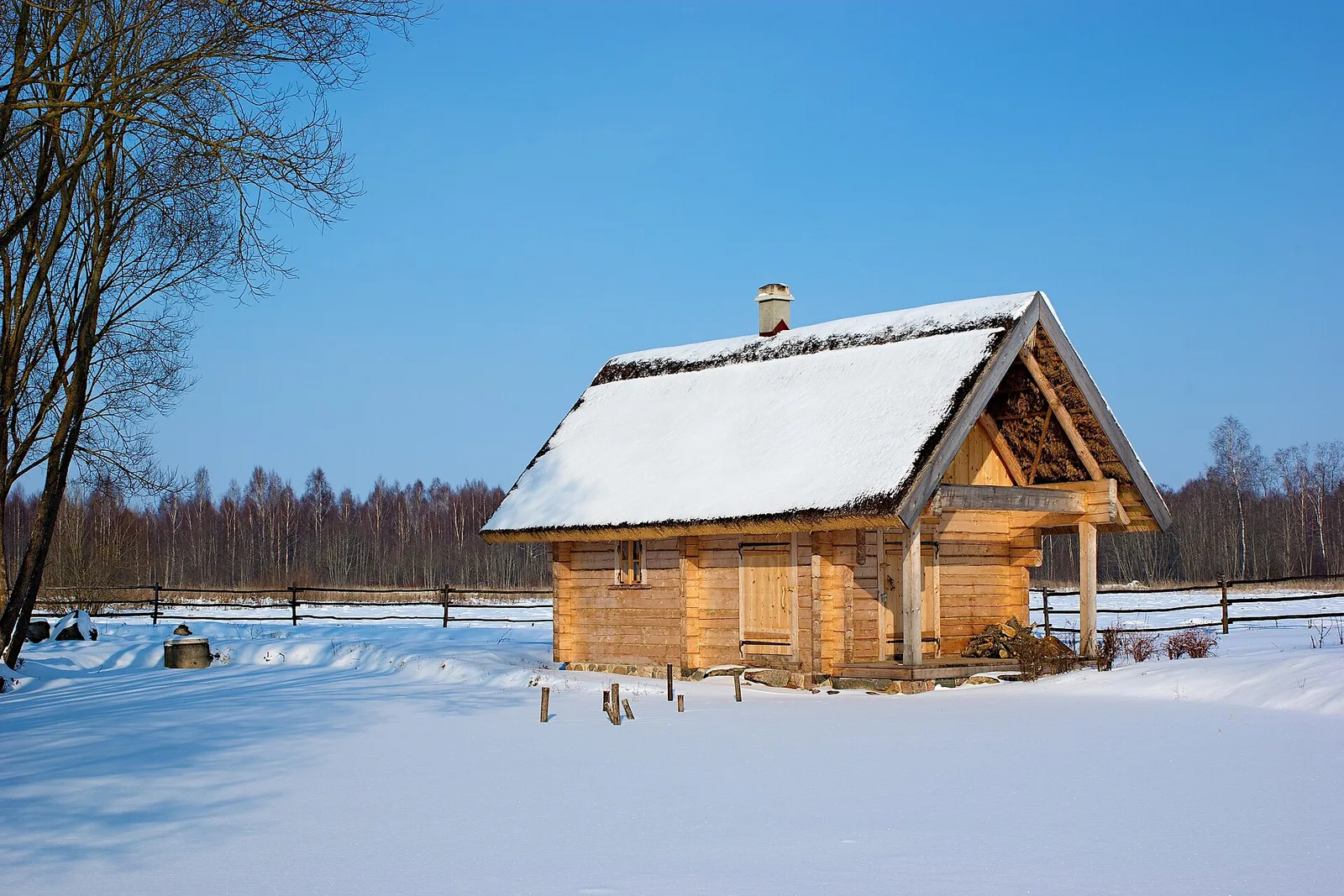 A snow-covered log sauna with a chimney and firewood stacked under its porch