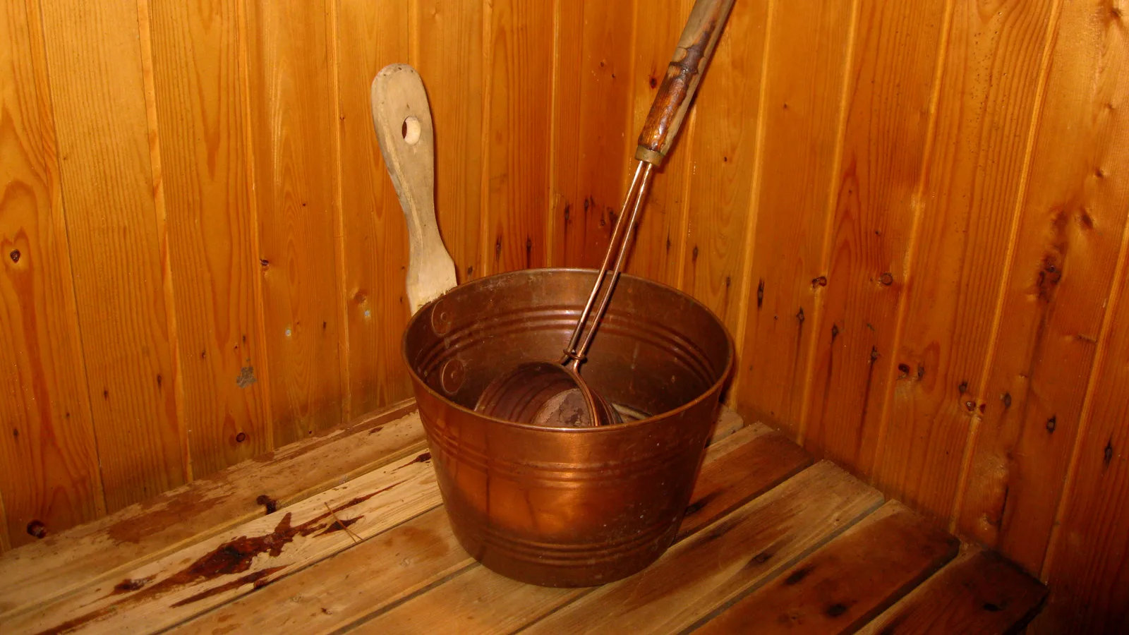 A copper sauna bucket with two ladles on the floor of a wooden Finnish sauna